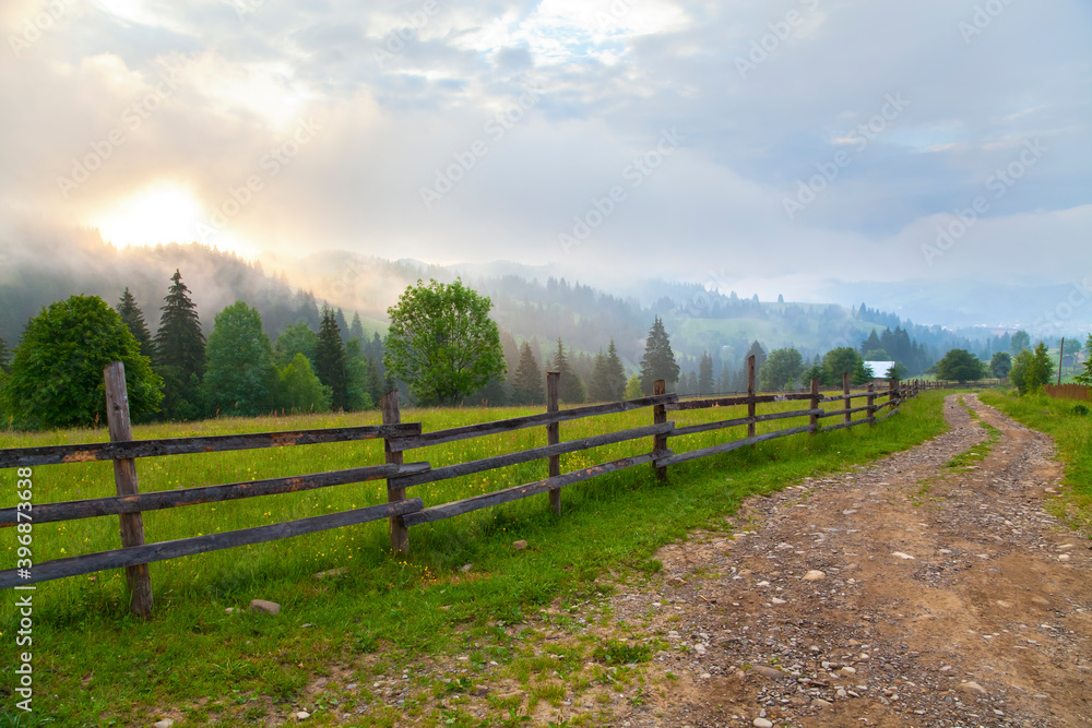 Road with  wooden fence along  the meadow, foggy mountains and morning sun. Ukraine, Verkhovyna.