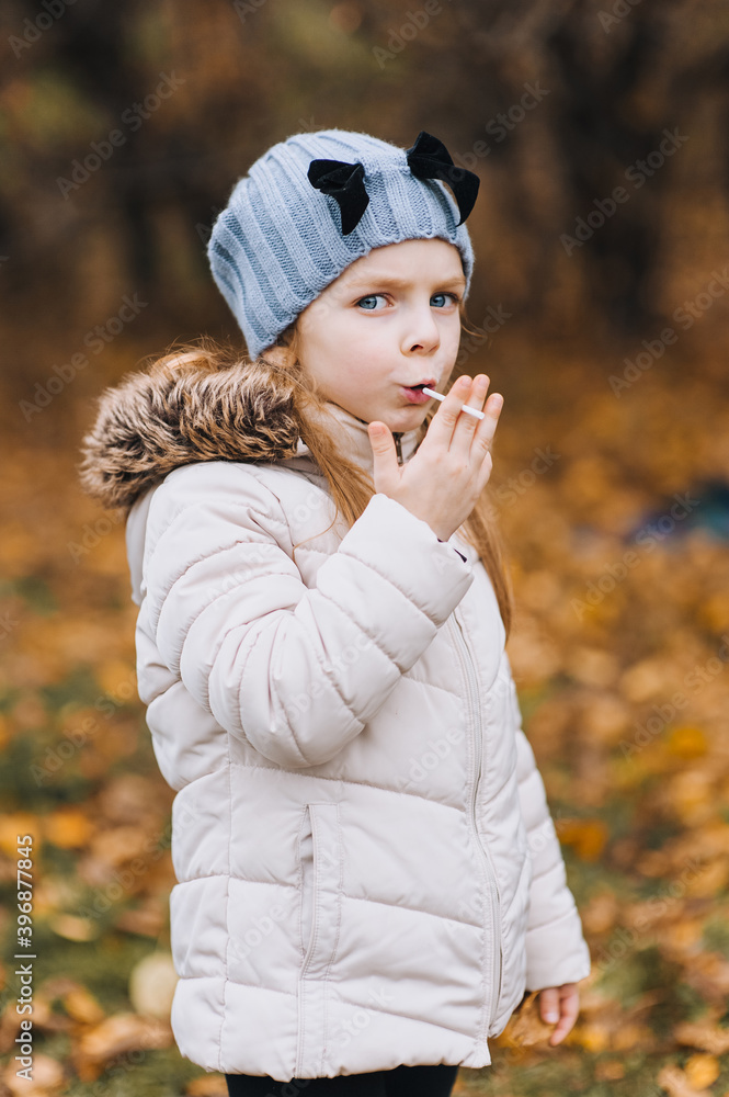 A beautiful little girl, a child in a jacket of preschool age, indulges, smokes a cigarette, imitating adults. Autumn funny portrait, photography. A beautiful little girl, a child in a jacket of preschool age, indulges, smokes a cigarette, imitating adults. Autumn funny portrait, photography.
