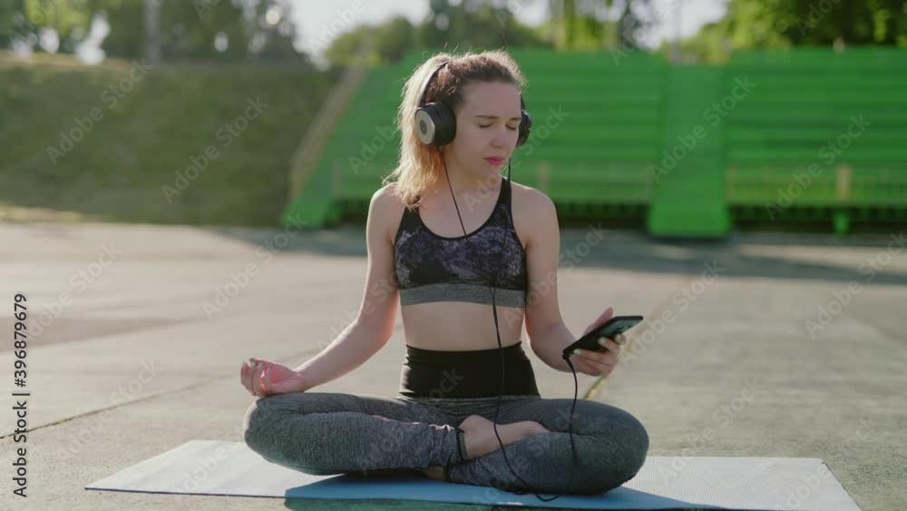 Relaxed young woman in sportswear meditating with music in headphones in lotus position in youga style. Female sitting with closed eyes in nature. Girl doing breathing exercises or practice. Harmony. Relaxed young woman in sportswear meditating with music in headphones in lotus position in youga style. Female sitting with closed eyes in nature. Girl doing breathing exercises or practice. Harmony.