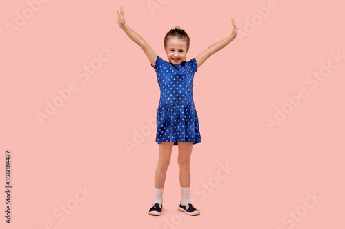 Happy child rejoices in victory hands raised up. The concept of winning. Human emotion. In a blue dress with a small star print. Pink isolated background.