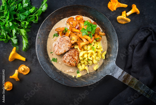 Modern style traditional fried pork filet medaillons in cream sauce with chanterelle mushrooms and spaetzle offered as top view on a rustic wrought iron skillet