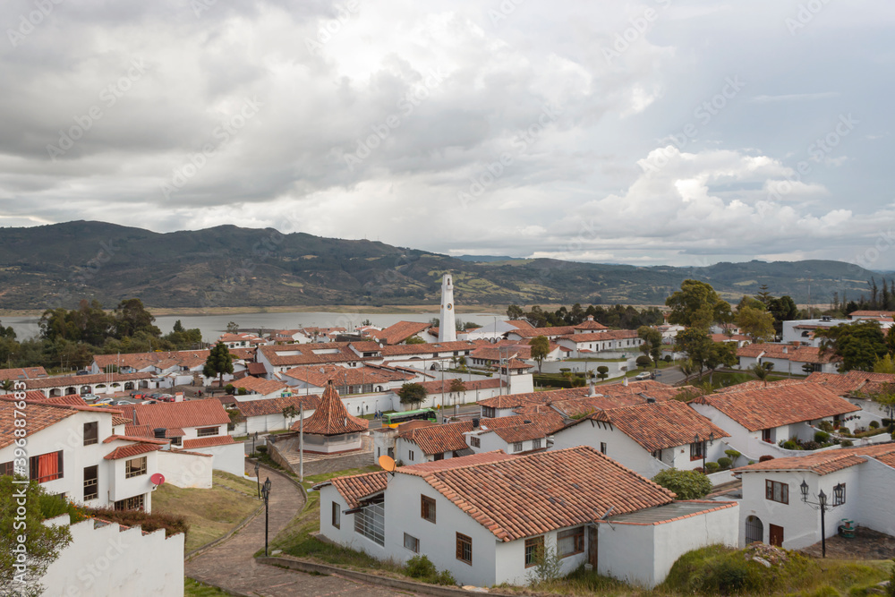 Fototapeta premium Guatavita colombian town cityscape with white structure, red ceramic roofs and andean mountains and tominé lake at background at evening. 