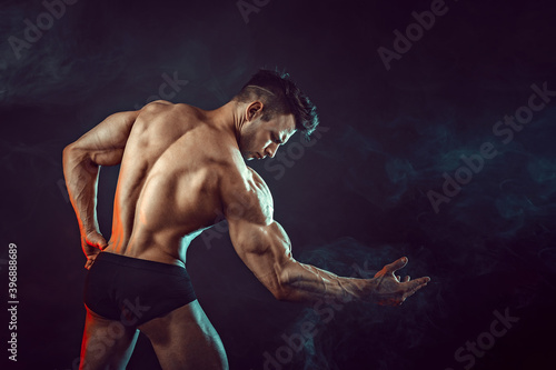 Athletic man flexing muscles in studio on dark background with smoke. Strong bodybuilder with perfect abs.