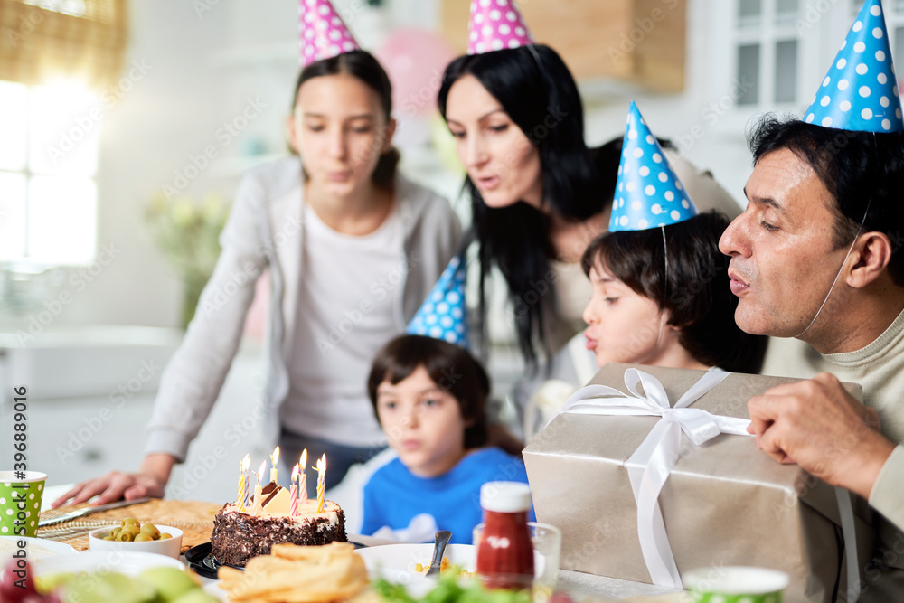 Joy. Close latin family with children wearing birthday caps, blowing candles on a cake while celebrating birthday together at home