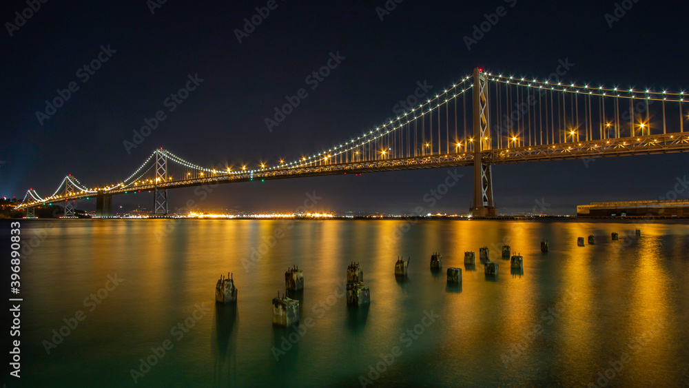 Fototapeta premium Night image of the Oakland Bridge as it leaves San Francisco and heads northwest into Oakland with lighted reflections of sunken pier ballasts