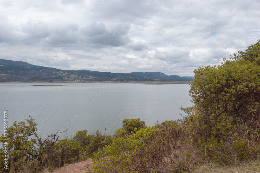 Guatavita tomine lake reservoir spring scene with white cloudy day, green trees and mountains at background