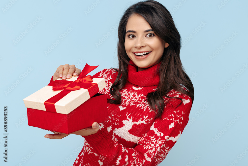 Surprised young brunette woman dressed in Christmas sweater opening gift box over blue background. Surprised young brunette woman dressed in Christmas sweater opening gift box over blue background.