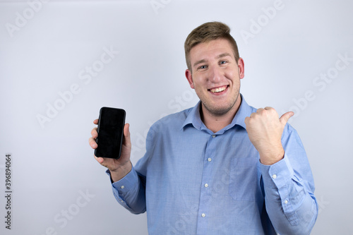 Business young man wearing a casual shirt over white background smiling and pointing to the copy space and holding a phone with the other hand