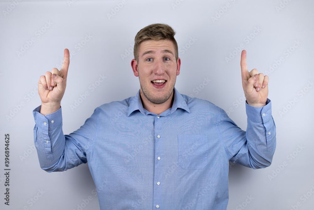 Business young man wearing a casual shirt over white background amazed and surprised looking at the camera and pointing up with fingers and raised arms Business young man wearing a casual shirt over white background amazed and surprised looking at the camera and pointing up with fingers and raised arms