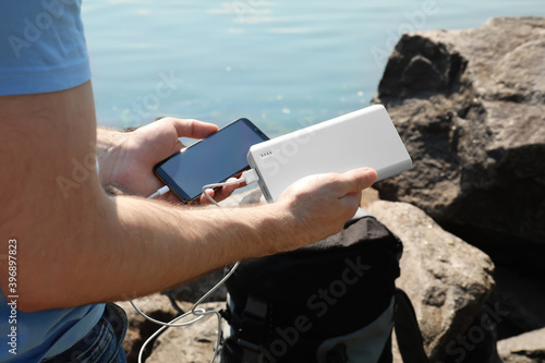 Man charging mobile phone with power bank on rocky mountain near river, closeup