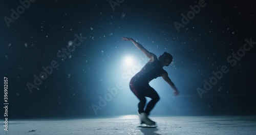 Cinematic shot of young female figure skater with headphones is performing training choreography on ice rink with snow falling. Concept of winter holidays, love and passion for sports and music. photo