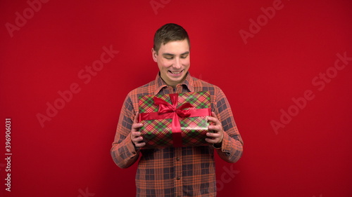 A young man shakes a Christmas present in his hands and rejoices. Shooting in the studio on a red background.
