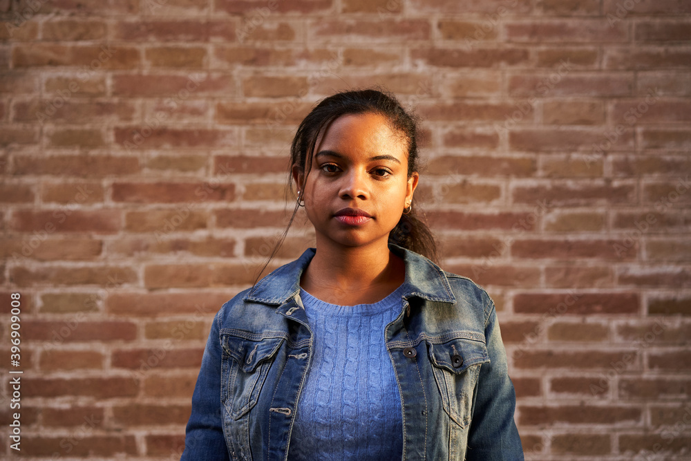 Portrait of young beautiful African American woman looking at camera with serious face outdoors. Serious millennial girl standing in front of a brick wall. People and emotions concept. Portrait of young beautiful African American woman looking at camera with serious face outdoors. Serious millennial girl standing in front of a brick wall. People and emotions concept.