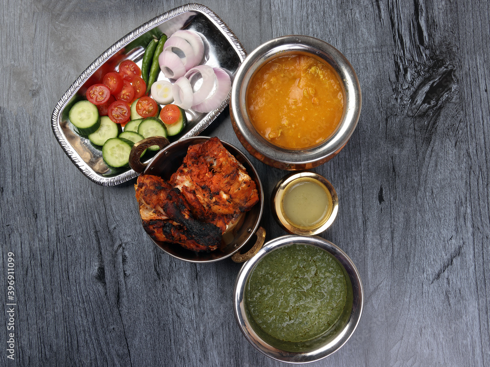 Spicy red Chicken grilled tikka tandoori nan bread green spinach curry yogurt sauce dal tomato cucumber onion salad set in metal stainless steel brass copper pot on black char wood background