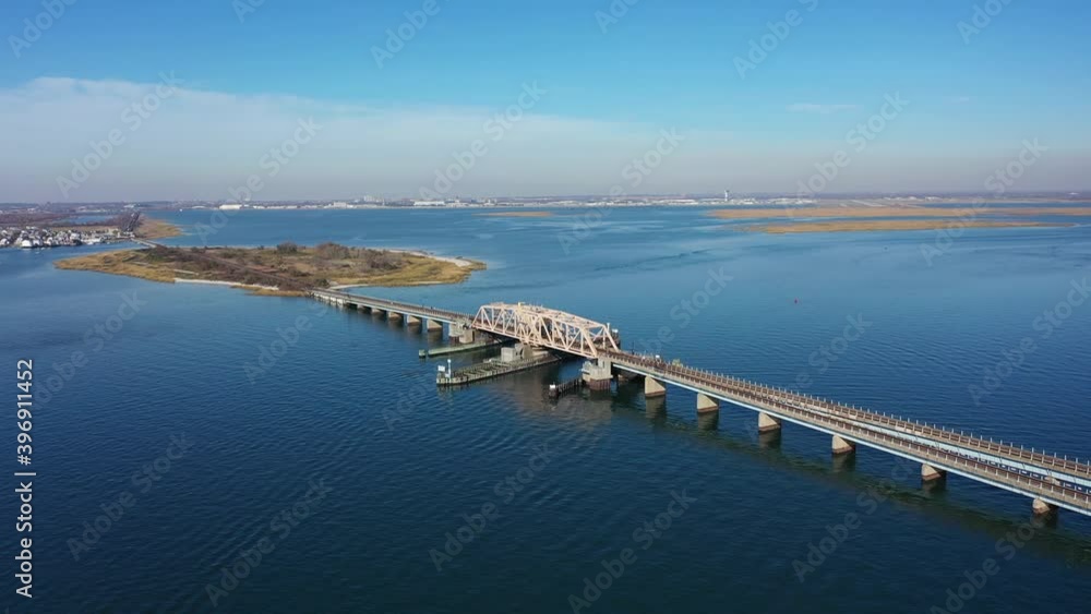 An aerial shot over an elevated train track with a swing bridge. The tracks crosses a bay in Queens, NY. The camera dolly out & pan right focusing on the bridge. It is a beautiful day. An aerial shot over an elevated train track with a swing bridge. The tracks crosses a bay in Queens, NY. The camera dolly out & pan right focusing on the bridge. It is a beautiful day.