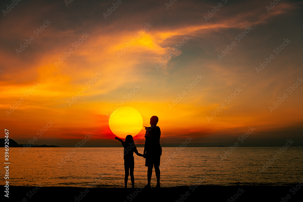 Silhouette young sister and brother stand on sand beach , hand point to see beautiful sun on sunset sky background.