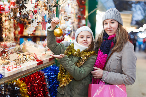 Smiling girl with woman are preparing for a Christmas and choosing balls on a tree outdoor. Focus on both persons