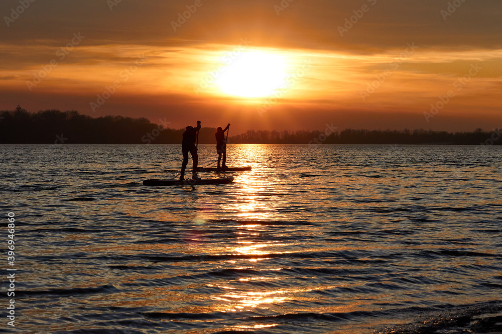 Silhouettes of a two boys rowing on a SUP (on stand up paddleboard) at sunset in a winter river