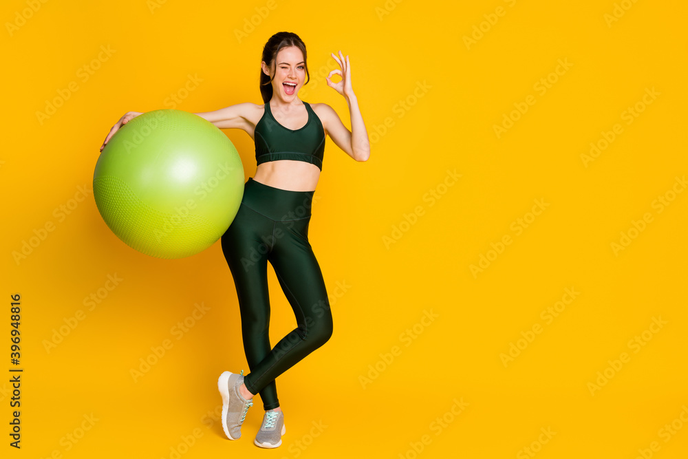 Full length body size view of nice cheerful girl holding in hands fitness ball showing ok-sign winking isolated over bright yellow color background.