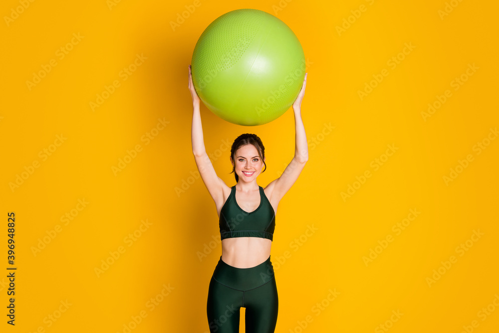 Portrait of nice attractive cheery girl holding in hands fitball over head doing pilates shaping isolated bright yellow color background Portrait of nice attractive cheery girl holding in hands fitball over head doing pilates shaping isolated bright yellow color background