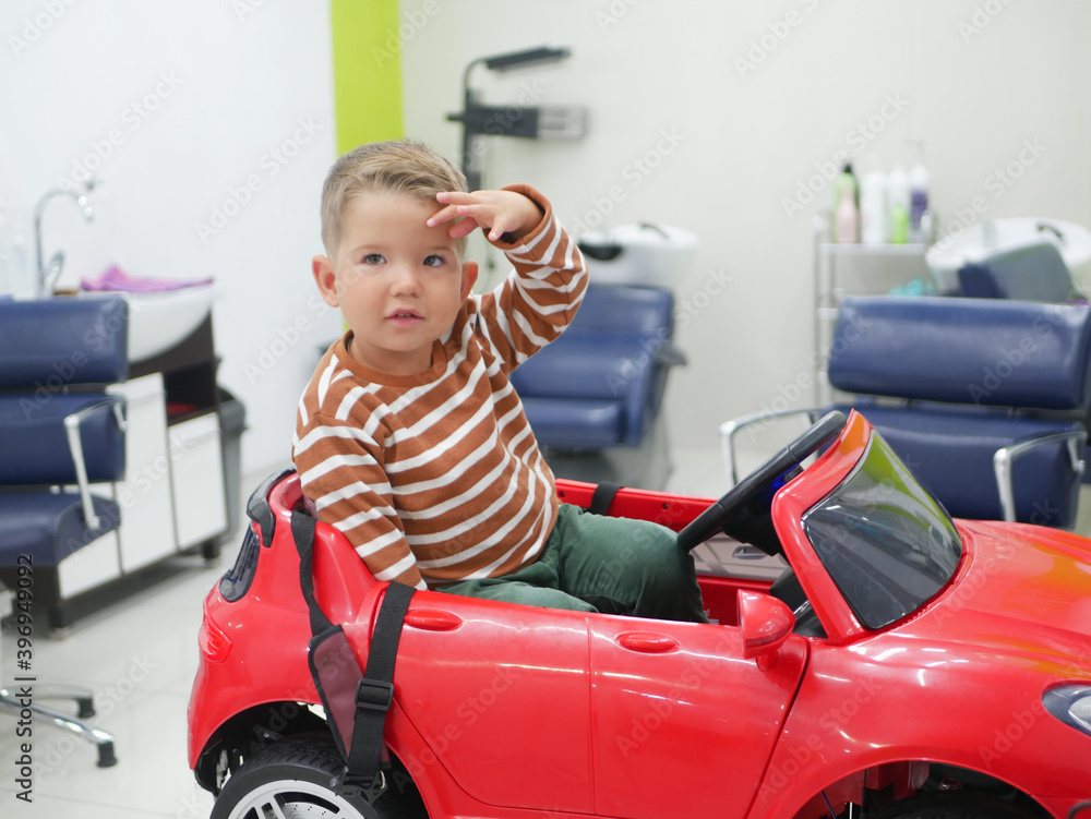 Smiling 2-3 years old toddler boy with new stylish haircut sitting in big toy car at the hairdresser. First haircut concept