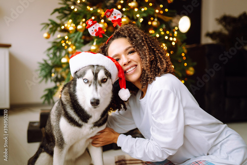 Beautiful woman playing and having fun with her dog while sitting near the christmas tree. Young lady hugging her dog with red christmas hat. Winter holidays. Light around. © maxbelchenko