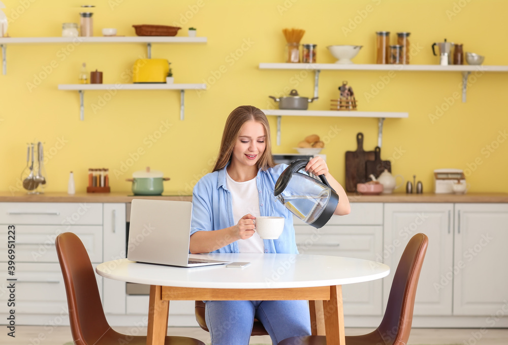 Young woman pouring boiled water from electric kettle into cup in kitchen Young woman pouring boiled water from electric kettle into cup in kitchen