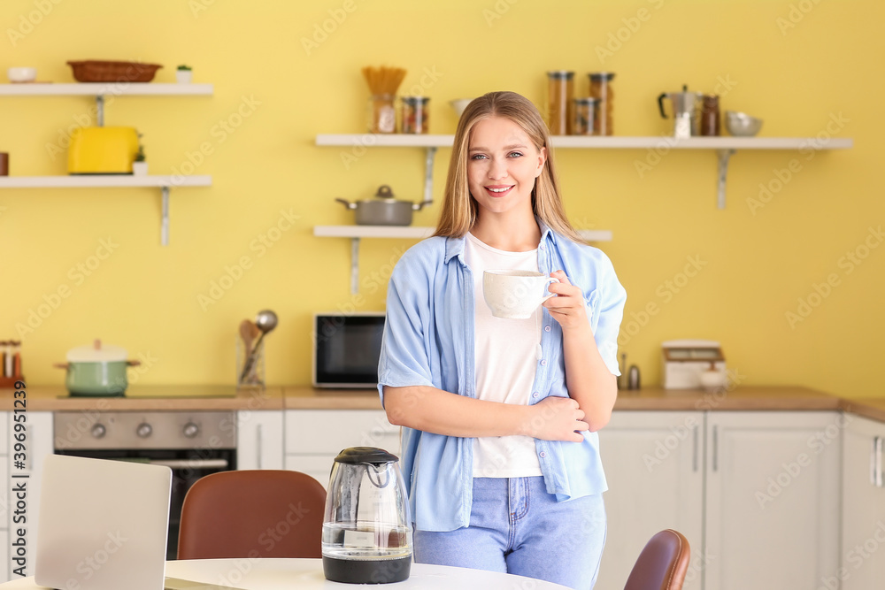 Young woman with cup and electric kettle on table in kitchen Young woman with cup and electric kettle on table in kitchen