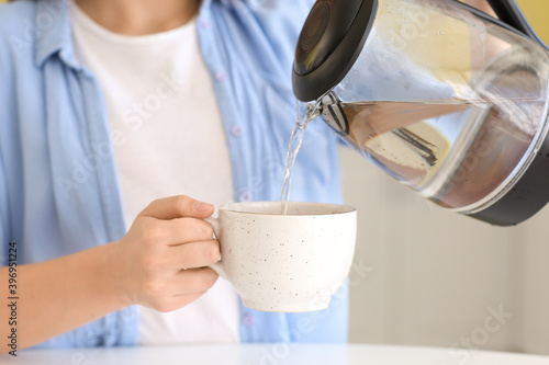 Young woman pouring boiled water from electric kettle into cup in kitchen, closeup