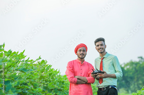 Young indian farmer holding remote in hand and control flying drone with agronomist at agriculture field