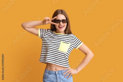 Beautiful woman with stylish sunglasses showing victory gesture on color background