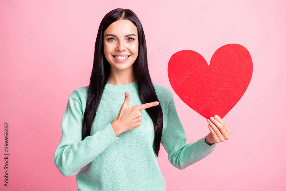 Photo portrait of woman pointing finger at big red heart card in one hand isolated on pastel pink colored background Photo portrait of woman pointing finger at big red heart card in one hand isolated on pastel pink colored background