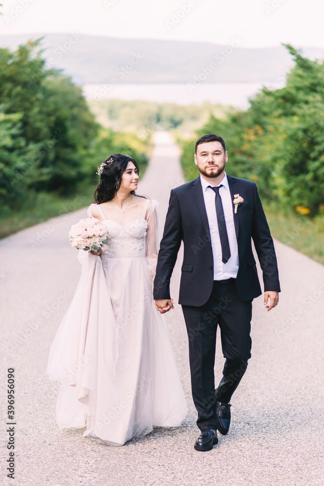 Naklejka premium Newlyweds groom in a stylish black suit, the bride in a powdery dress, a photo session in nature, the road against the background of mountains and a reservoir in summer on a sunny day