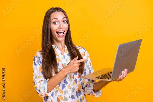 Photo portrait of excited girl with open mouth pointing finger at laptop in hand isolated on vivid yellow colored background