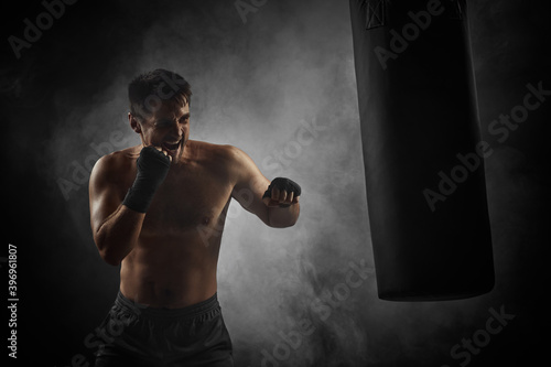 Aggressive boxer in black boxing wraps punching in boxing bag on dark background with smoke © producer