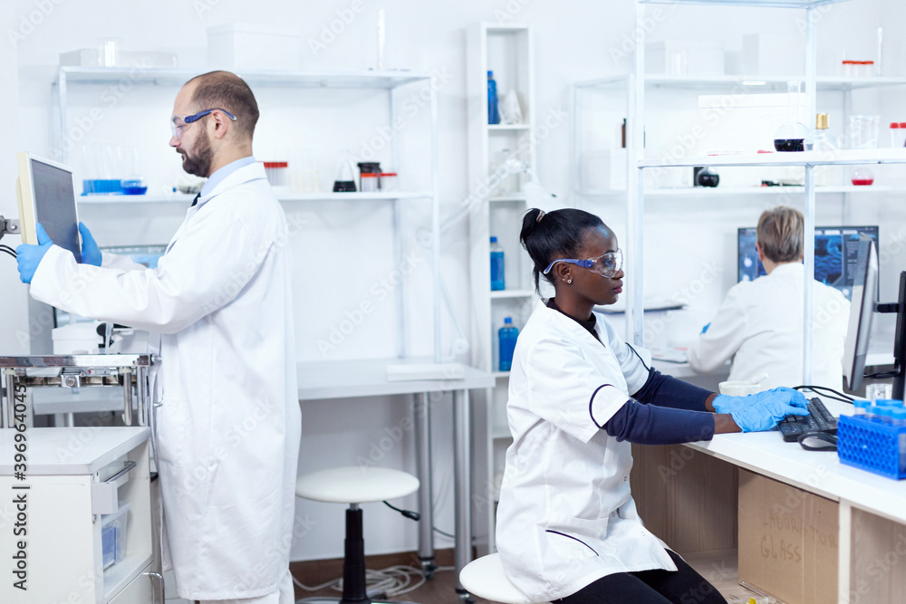 Obraz premium African scientist working on computer wearing protection gear doing analysis. Multiethnic team of researchers working in microbiology lab testing solution for medical purpose.