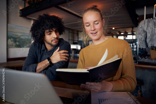 Young male and female business people looking at notes in diary while working using laptop in modern cafe