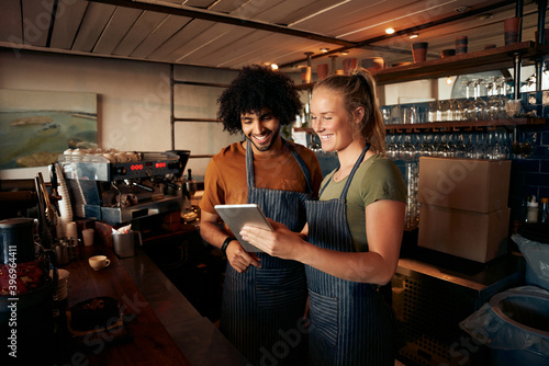 Smiling waiter and waitress wearing apron using digital tablet standing behind counter in cafe