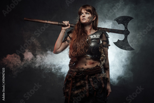 Beautiful and at the same time barbaric female warrior from nord poses in dark foggy background holding two handed axe.