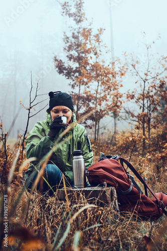 Woman with backpack having break during autumn trip drinking a hot drink from thermos flask on autumn cold day. Active middle age woman wandering around a forest actively spending time