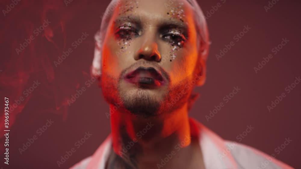 Portrait of handsome man with white hair and bold make-up with glitter blowing smoke and looking at camera in red lighting Portrait of handsome man with white hair and bold make-up with glitter blowing smoke and looking at camera in red lighting