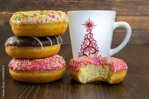 Glazed donuts of various colors, a bitten off donut and a cup of hot coffee, close-up on a dark background.