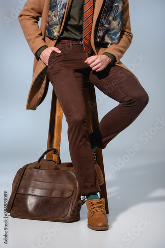 cropped view of stylish man in winter coat sitting on wooden chair with hand in pocket near briefcase on grey