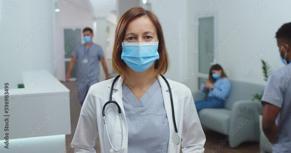 Close up portrait of beautiful Caucasian female doctor wearing medical mask standing in clinic hall and looking at camera. Mixed-races healthcare workers on background. Covid 19 coronavirus concept Close up portrait of beautiful Caucasian female doctor wearing medical mask standing in clinic hall and looking at camera. Mixed-races healthcare workers on background. Covid 19 coronavirus concept