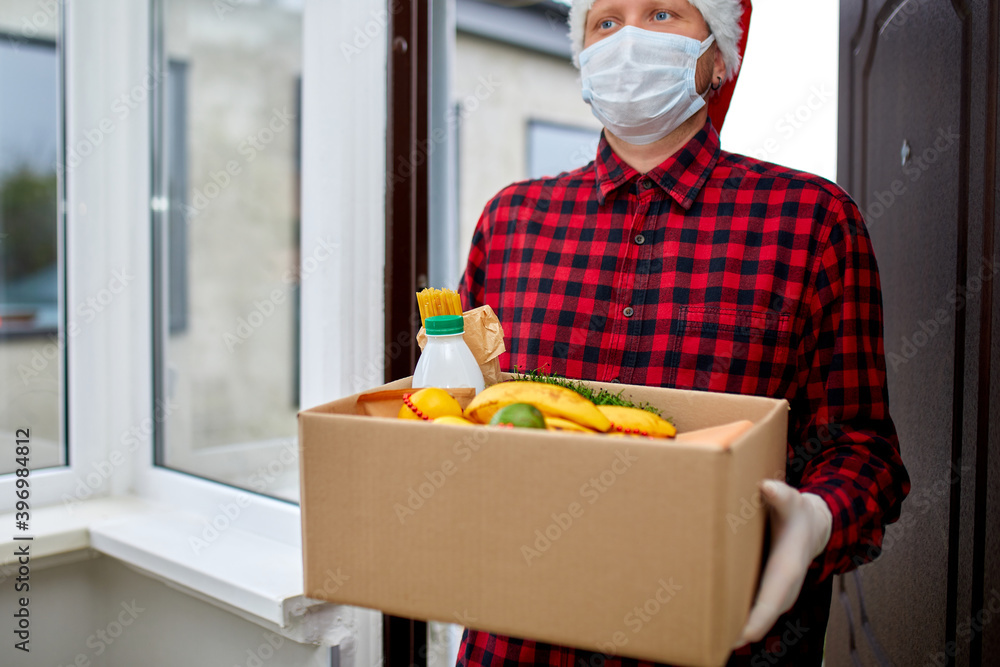 Volunteer man in Santa hat and protective mask and gloves delivery donation box at home in Christmas Volunteer man in Santa hat and protective mask and gloves delivery donation box at home in Christmas