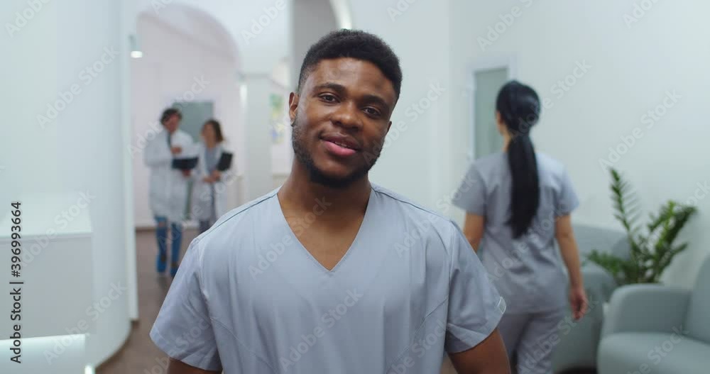 Close up portrait of African American young joyful male doctor surgeon standing in hospital and looking at camera in good mood. Caucasian healthcare professionals on background. Medic concept Close up portrait of African American young joyful male doctor surgeon standing in hospital and looking at camera in good mood. Caucasian healthcare professionals on background. Medic concept