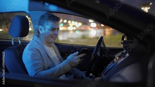 Side shot portrait of man in electric car typing on smartphone and smiling to camera. Handsome happy male sitting in vehicle in good mood and texting on mobile phone. Transport concept