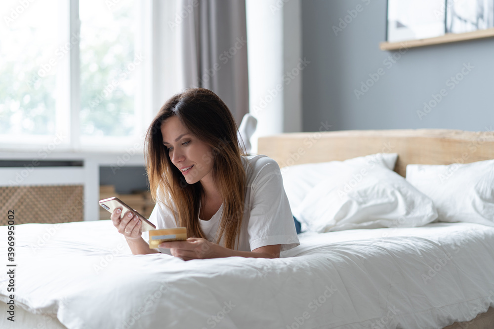 Beautiful young woman relaxing on bed at home, using mobile phone, holding credit card, shopping. Beautiful young woman relaxing on bed at home, using mobile phone, holding credit card, shopping.