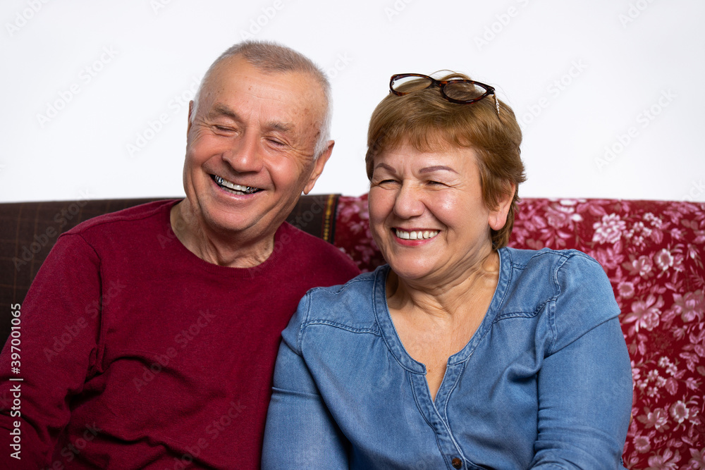 An elderly smiling married couple looking at the camera, sitting home on couch. An elderly smiling married couple looking at the camera, sitting home on couch.