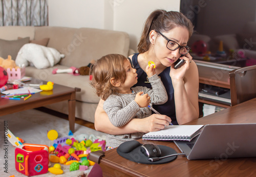 Young beautiful business woman talking on mobile phone and working on a laptop. Mother playing with child during working. Women powerful. Lady working at home during quarantine because of coronavirus.